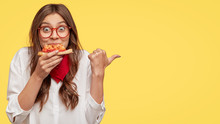 Lunch Time Concept. Attractive Cheerful Woman Eats Delicious Italian Pizza, Surprised With Wonderful Taste, Indicates With Thumb Aside, Shows Where Pizzeria Situated, Suggests To Visit And Have Snack
