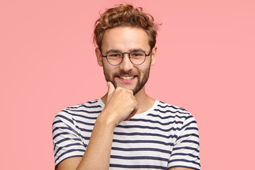 Wall Mural - Horizontal shot of satisfied self assured guy with pleasant smile, keeps hand on chin, wears casual clothes, rejoices good resuls or achievement, stands against pink background. Happiness concept