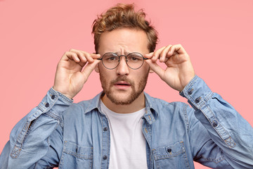 Wall Mural - Horizontal shot of pleasant looking guy looks scrupulously through glasses, tries to notice something, has bad sight, dressed in fashionable shirt, poses against pink studio wall. Facial expressions