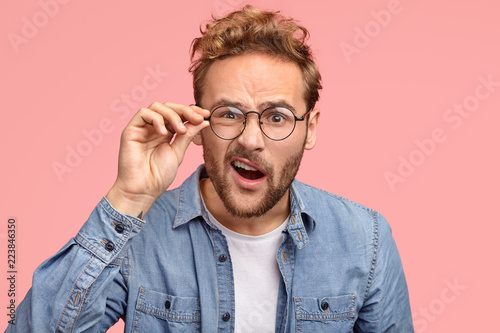 Indignant young Caucasian guy looks desperately at camera, wears  spectacles, opens mouth, has surprised gaze, expresses frustration, wears  round spectacles, stands against pink studio background - Buy this stock  photo and explore