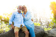 Elderly couple sitting on a rock they are in love and happiness and embracing with smiling in the park with sunshine as a background.