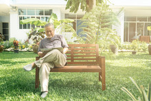 Old Man Sitting On Bench In Garden Reading Newspaper.