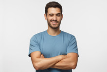 Handsome Young Man In Blue T-shirt, With Crossed Arms Smiling And Winking, Looking At Camera Isolated On Gray Background