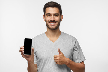 Wall Mural - attractive happy young man holding blank smartphone, smiling at camera, showing thubms up gesture, i