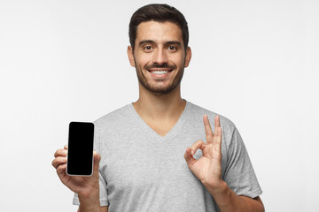 Wall Mural - smiling young man holding blank smartphone with empty black sreen, showing ok or okay gesture with o
