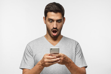 Close up portrait of young man in gray t-shirt, holding smartphone, looking scared and confused reading text message or e-mail