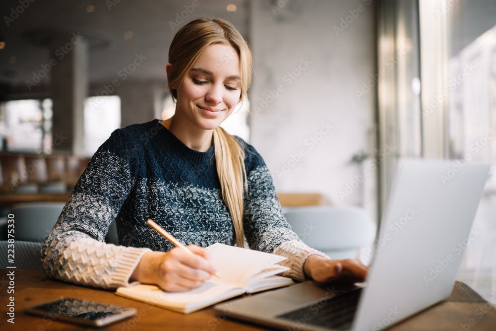 Young attractive university student using laptop computer, studying at modern library. Cheerful caucasian woman writes notes, planning working process, sitting in loft cafe. Exam preparation.  - obrazy, fototapety, plakaty 