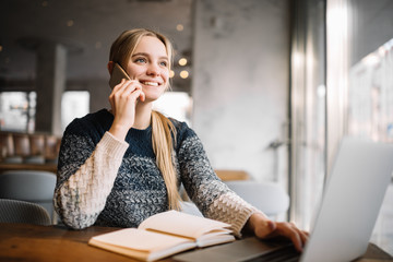 Young cheerful  woman with emotional face speaks on mobile phone and smiling in loft office. Positive freelancer using laptop computer and smartphone at workplace. Startup. Successful business.