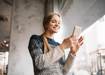 Smiling, positive blonde female  holding smartphone and booking travel online in modern coffee shop. Hipster woman with beautiful face downloading application. Mobile banking. Cash back. 