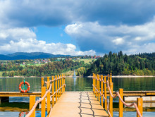 Pier To The Lake With Lifebuoy, Mountains And Cloudy Sky, View Of The Sailboat And The Village