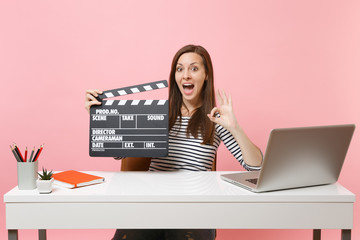 Wall Mural - Amazed woman showing OK sign holding classic black film making clapperboard and working on project while sit at office with laptop isolated on pink background. Achievement business career. Copy space.