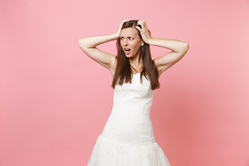 Portrait of concerned shocked bride woman in white wedding dress standing looking aside clinging to head isolated on pink pastel background. Wedding celebration concept. Copy space for advertisement.