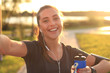 Young attractive woman in sports clothing looking at phone and smiling while taking selfie, during outdoors workout.