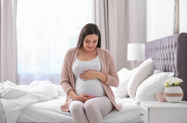 Poster - Young beautiful pregnant woman sitting on bed and touching her belly at home