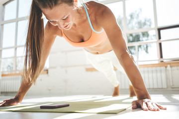 Sporty strong woman doing push-ups on fitness mat and looks on smartphone for counts results. Female athlete exercising push up in sunny gym. Toned color