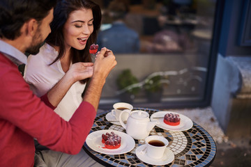 So delicious. Loving young couple sitting at the table with cups of tea and teapot. Gentleman giving raspberry cake to smiling lady