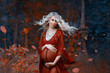 © kharchenkoirina - a beautiful pregnant woman with blond hair in long red dress and shiny necklace is standing in the forest, gently smiling into the camera, curly blonde hair fluttering in the wind. autumn art photo.