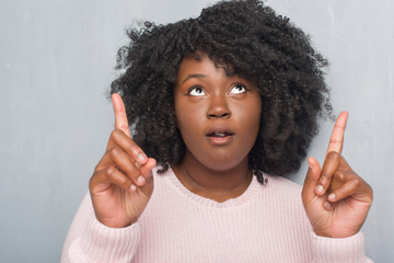 Canvas Print - Young african american plus size woman over grey grunge wall wearing winter sweater amazed and surprised looking up and pointing with fingers and raised arms.