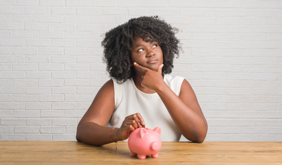 Poster - Young african american woman sitting on the table holding piggy bank serious face thinking about question, very confused idea