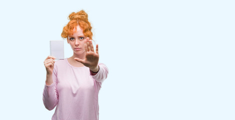 Sticker - Young redhead woman holding blank notebook with open hand doing stop sign with serious and confident expression, defense gesture