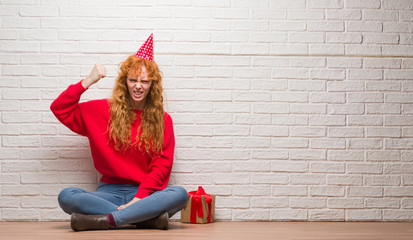 Wall Mural - Young redhead woman sitting over brick wall wearing birthday hat annoyed and frustrated shouting with anger, crazy and yelling with raised hand, anger concept