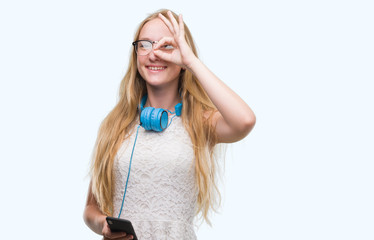 Canvas Print - Blonde teenager woman holding smartphone and wearing headphones with happy face smiling doing ok sign with hand on eye looking through fingers