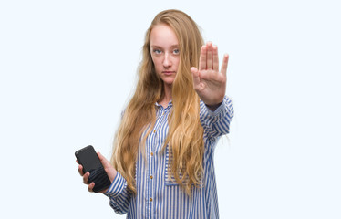 Poster - Blonde teenager woman using smartphone with open hand doing stop sign with serious and confident expression, defense gesture