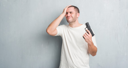 Young caucasian criminal man over grey grunge wall holding gun stressed with hand on head, shocked with shame and surprise face, angry and frustrated. Fear and upset for mistake.