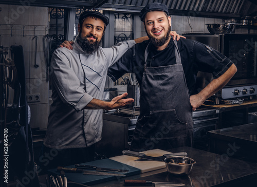 Fotografía Two happy bearded cooks friend dressed in uniforms and hats embrace while posing in kitchen