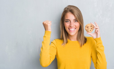 Wall Mural - Beautiful young woman over grunge grey wall eating chocolate chip cooky screaming proud and celebrating victory and success very excited, cheering emotion