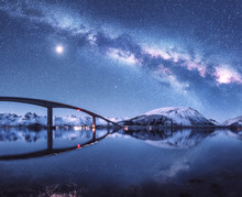 Bridge And Starry Sky With Milky Way Over Snow Covered Mountains Reflected In Water. Night Landscape With Road, Snowy Rocks, Sky With Moon, Milky Way, Stars, Sea. Winter In Lofoten Islands, Norway