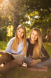 © Sunrise Team - Young beautiful ladies students sitting in the park outdoors on grass holding copybook doing homework.