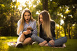 © Sunrise Team - Shocked surprised girls sisters students sitting in the park outdoors on grass using mobile phone listening music with earphones.