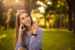 © Sunrise Team - Happy cheerful smiling girl student sitting in the park outdoors on grass have a rest talking by mobile phone pointing to copyspace.