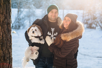 Cute young couple having fun in winter forest with their pretty little white dog. Man and woman in sweater with a snowflake