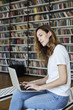 Portrait of smiling smart woman model with opened laptop in a library, bookshelf behind, long hair. Hipster college student lady.
