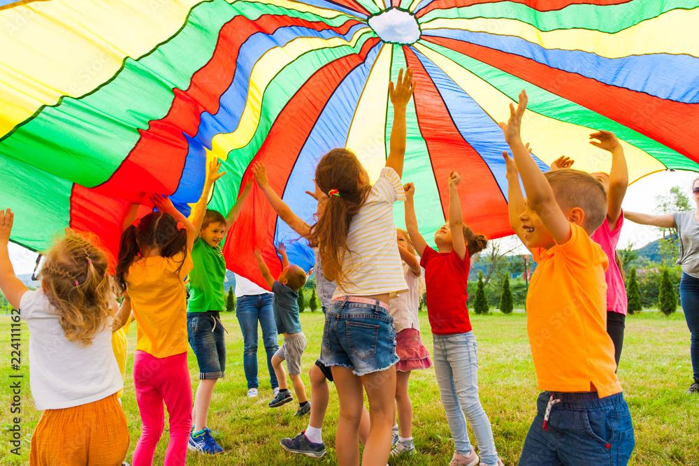 Joyous classmates jumping under colorful parachute in the summer outdoors - obrazy, fototapety, plakaty 