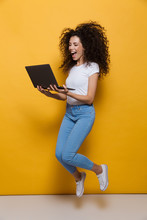 Full Length Photo Of Funny Woman 20s Wearing Casual Clothes Smiling And Jumping While Holding Black Laptop, Isolated Over Yellow Background