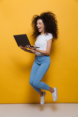 Poster - Full length photo of funny woman 20s wearing casual clothes smiling and jumping while holding black laptop, isolated over yellow background