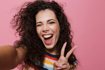 Wall Mural - Image of young woman 20s with curly hair laughing and taking selfie photo, isolated over pink background