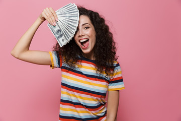 Poster - Image of surprised woman 20s with curly hair holding fan of dollar money, isolated over pink background