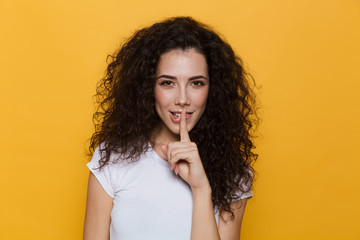 Wall Mural - Image of charming woman 20s with curly hair holding finger at lips, isolated over yellow background