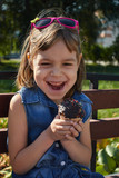 Fototapeta Kwiaty - A girl with blond hair sits on a park bench and eats a muffin.
