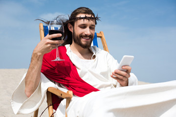 smiling Jesus resting on sun lounger with glass of wine and using smartphone in desert