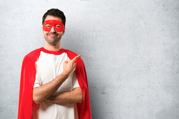 Wall Mural - Superhero man with mask and red cape pointing to the side with a finger to present a product or an idea while looking forward smiling on textured grey background