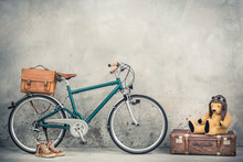 Retro Bicycle With Leather Mailman's Bag, Old Sneakers And Teddy Bear Toy In Leather Aviator's Hat And Goggles Sitting On Aged Classic Travel Suitcase Front Concrete Wall. Vintage Style Filtered Photo