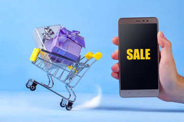 Gifts in a shopping cart and mobile phone on a blue background. Online shopping, shopping concept. Discounts and sales