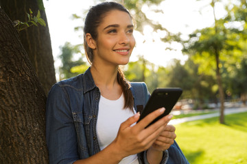 Sticker - Beautiful woman student walking in park looking aside using mobile phone.