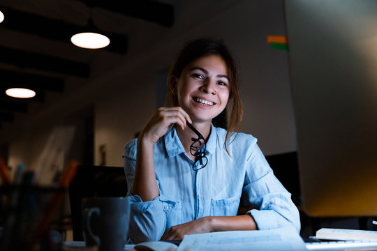 Cheerful young woman graphic designer using pc computer working at night in office looking camera.