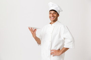 Cheerful handsome young chef man standing isolated over white wall background holding plate.